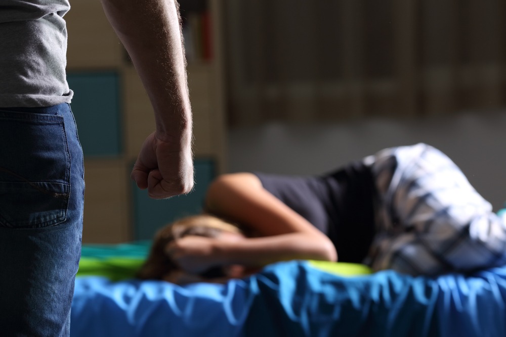 A mistreatment victim lies on a bed with her abuser standing over her.