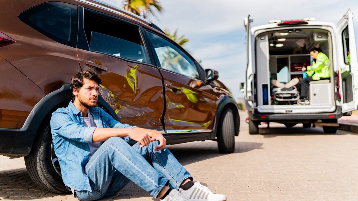 man-sitting-on-the-ground-against-his-car-after-an-accident-with-ambulance-nearby