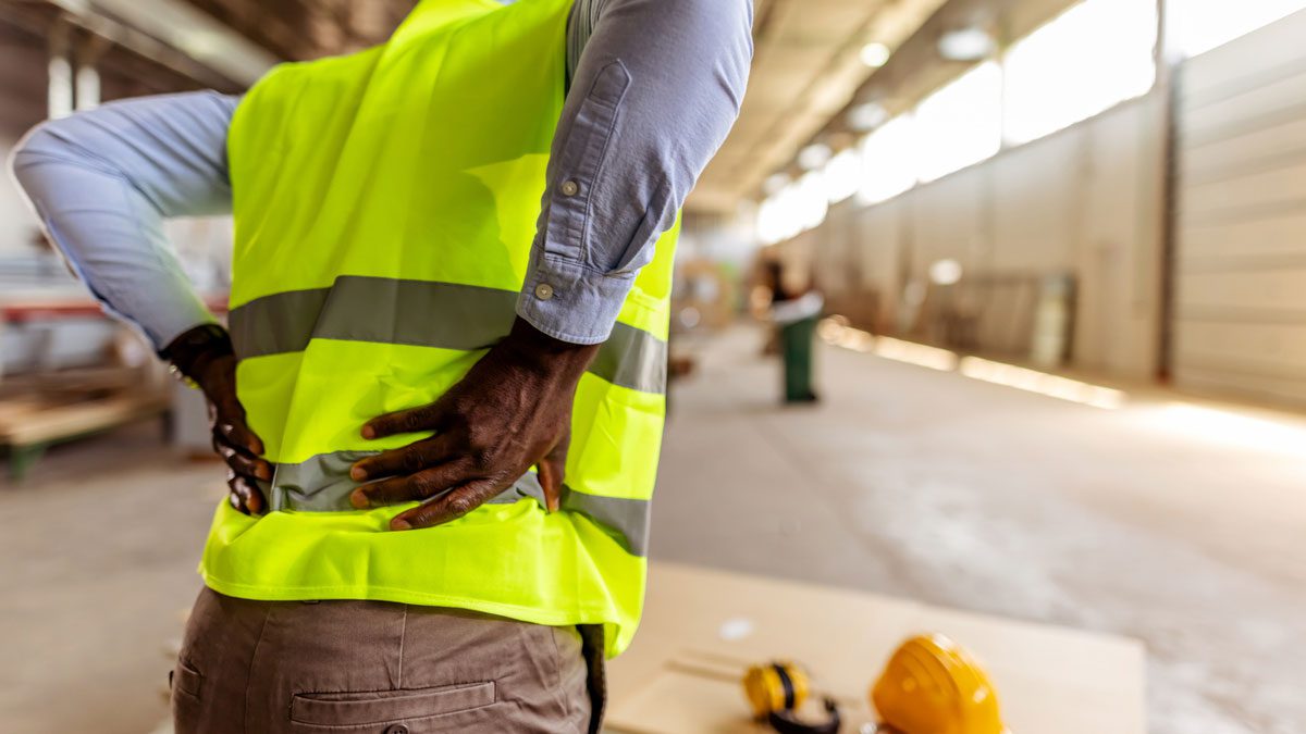 A factory worker in a high-visibility vest holding his back to indicate pain