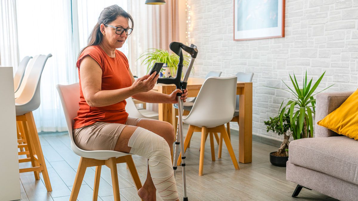 woman with leg in bandage sitting on chair holding crutches and using phone